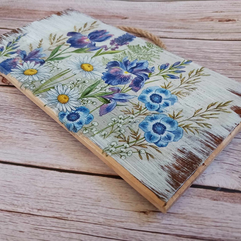 May include: A wooden sign with a distressed white paint finish features a floral design with blue and purple flowers. The sign has a rope hanger.