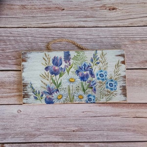 May include: A wooden sign with a white background and a floral design featuring blue and purple irises, white daisies, and blue wildflowers.