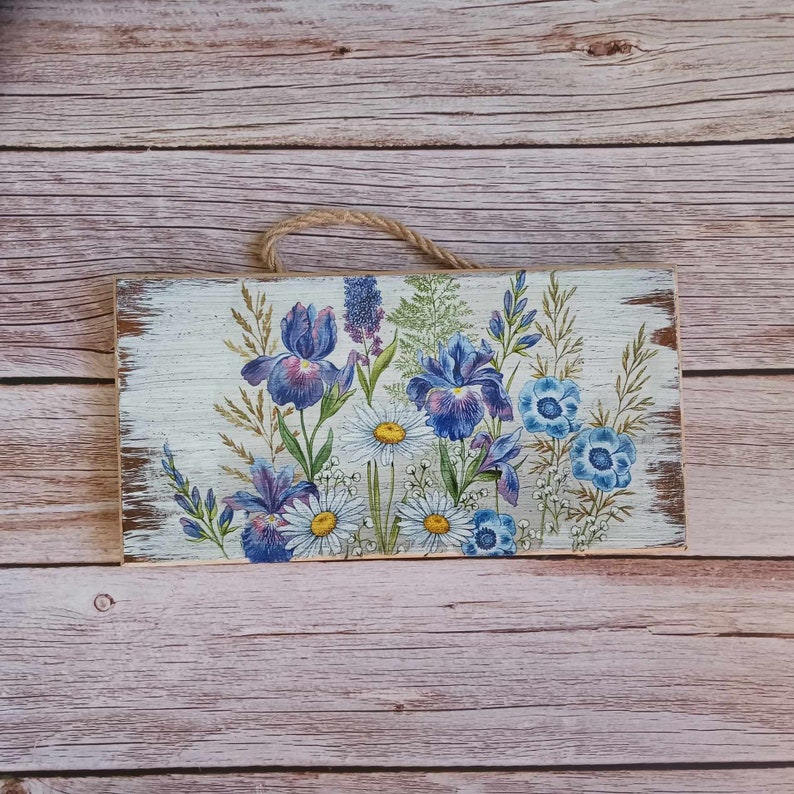 May include: A wooden sign with a white background and a floral design featuring blue and purple irises, white daisies, and blue wildflowers.