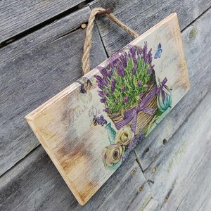 May include: A wooden sign with a distressed whitewash finish features a floral design with purple lavender flowers in a wicker basket. The sign has the text "Fleur De" and "Lavender" printed on it.