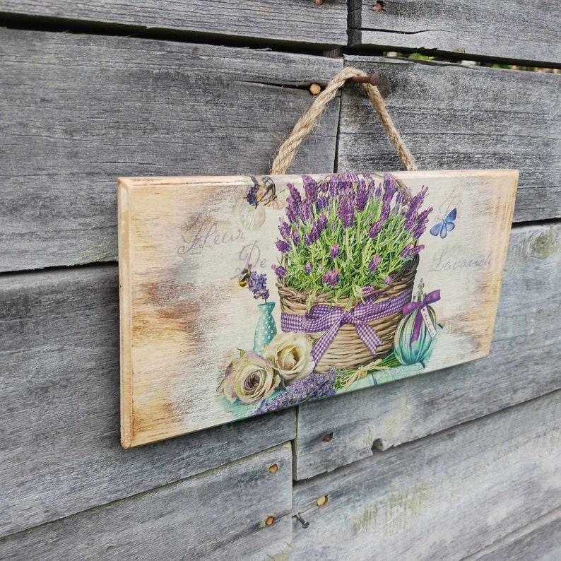May include: A wooden sign with a lavender floral design. The sign features a basket of lavender flowers, a butterfly, and the text "Fleur De Lavande".