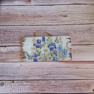May include: A wooden sign with a painted floral design featuring purple irises and white daisies. The sign has a distressed white background and is hung with twine.