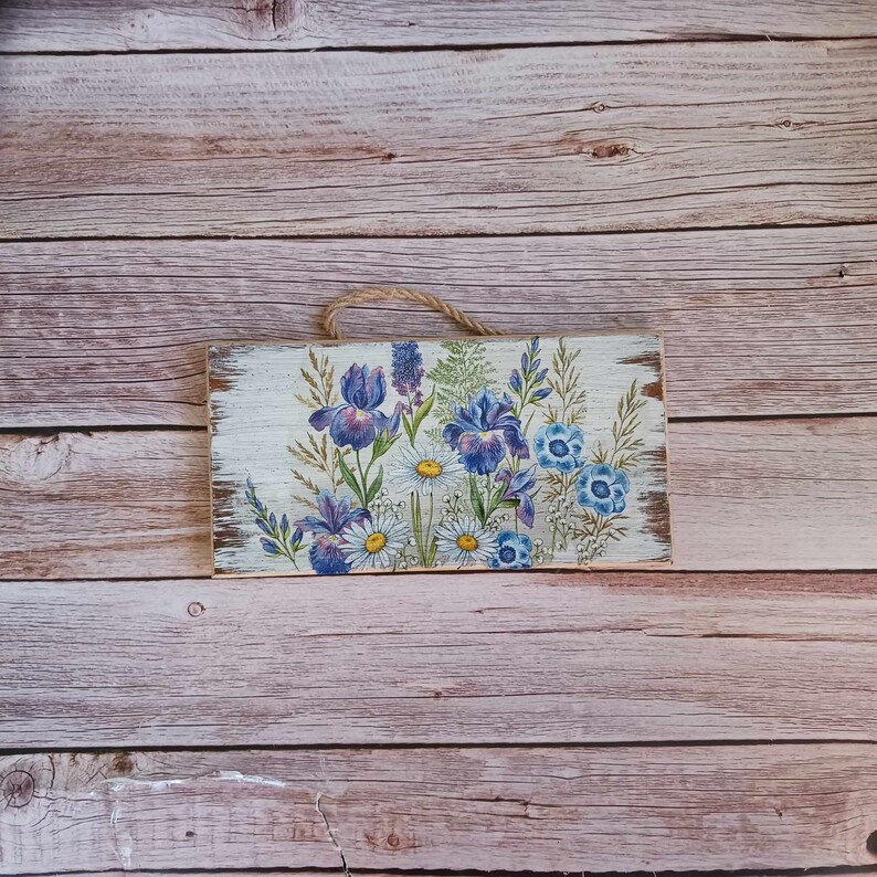 May include: A wooden sign with a painted floral design featuring purple irises and white daisies. The sign has a distressed white background and is hung with twine.