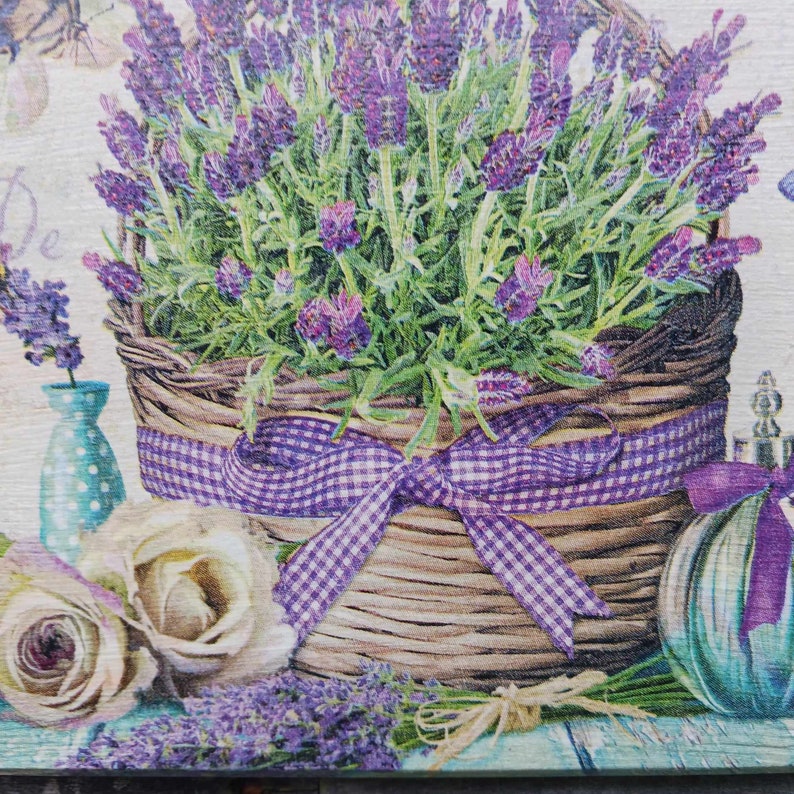 May include: A wicker basket filled with purple lavender flowers, tied with a purple and white gingham ribbon. The basket is sitting on a wooden surface with two white roses and a blue glass bottle.