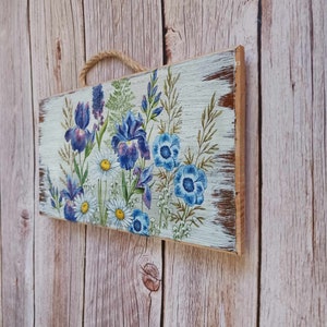 May include: A wooden sign with a distressed white paint finish features a floral design with blue and purple irises and white daisies. The sign has a rustic, vintage look.