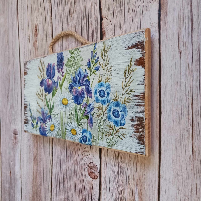 May include: A wooden sign with a distressed white paint finish features a floral design with blue and purple irises and white daisies. The sign has a rustic, vintage look.