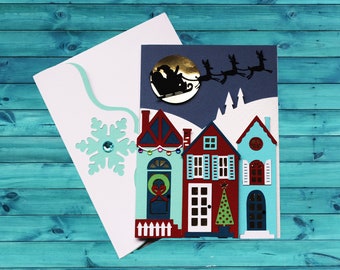 May include: A handmade Christmas card featuring a winter village scene with three colorful houses. Santa Claus in his sleigh flies across a dark blue sky. The card includes a white envelope with a snowflake embellishment.