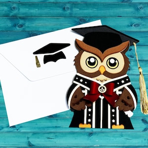 May include: A graduation-themed card featuring a cartoon owl wearing a graduation cap and gown. The owl is brown, with large eyes and a red bow tie. A white envelope with a graduation cap cutout is also present. The card has a gold tassel.