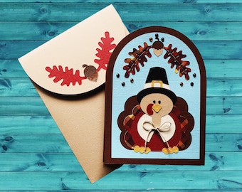 May include: A handmade greeting card featuring a layered paper turkey wearing a black pilgrim hat, positioned next to a matching cream-colored envelope decorated with red paper oak leaves and a brown paper acorn cutout on the envelope flap.
