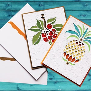 May include: Three white cards with orange and brown borders. The cards feature cut-out designs of a pineapple, a berry branch, and a floral pattern. The cards are on a blue wooden background.