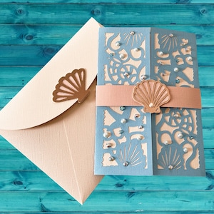 May include: A light blue and cream-colored invitation suite with a seashell theme. The invitation features laser-cut details, a peach-colored belly band, and pearl accents. An envelope with a fan-shaped embellishment completes the set.