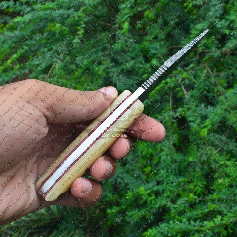 May include: A close-up of a knife with a white and red handle. The blade is made of Damascus steel and has a textured finish. The handle is made of bone and has a red stripe running down the center. The knife is being held in a hand.