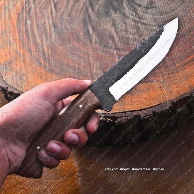 May include: A hand holds a custom-made knife with a dark brown wooden handle and a black blade. The blade has a white edge and is slightly curved. The knife is resting on a wooden surface.