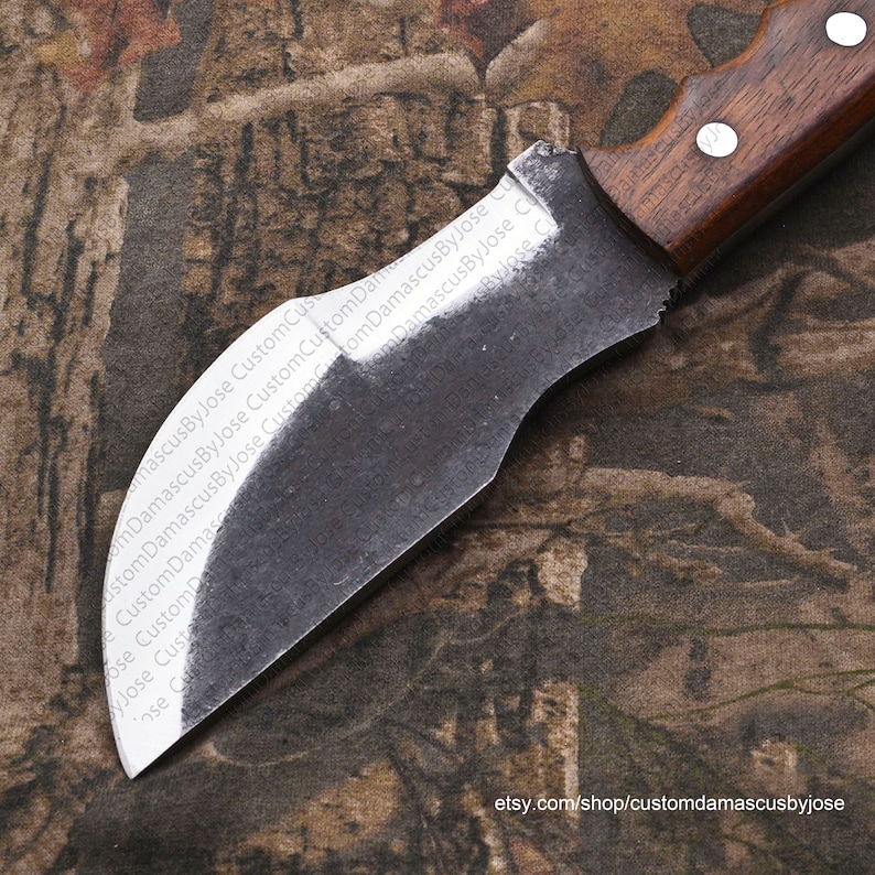 Puede incluir: Un cuchillo de acero damasco hecho a mano, curvo, con mango de madera. La hoja est&aacute; parcialmente pulida y tiene una ligera curva. El mango es de madera marr&oacute;n con un ligero patr&oacute;n de veta. El cuchillo est&aacute; sobre un fondo de tela de camuflaje.