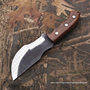 Puede incluir: Un cuchillo hecho a mano con un mango de madera marr&oacute;n oscuro y una hoja de acero gris curvada. El cuchillo est&aacute; apoyado sobre un fondo de tela de camuflaje.