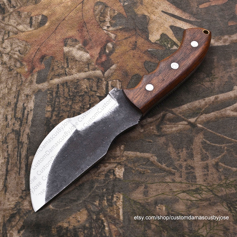 Puede incluir: Un cuchillo hecho a mano con un mango de madera marr&oacute;n oscuro y una hoja de acero gris curvada. El cuchillo est&aacute; apoyado sobre un fondo de tela de camuflaje.