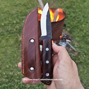 Könnte beinhalten: Ein handgemachtes Damaszener-Messer mit einem braunen Holzgriff und einer Lederscheide. Das Messer ist scharf und hat eine spitze Spitze. Das Messer ist scharf, und der Hintergrund ist verschwommen.