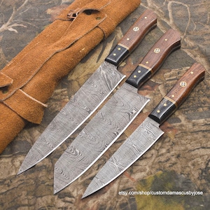 Könnte beinhalten: Set aus drei Damastmesser mit Holzgriffen und einer Lederrolle zur Aufbewahrung. Die Messer haben eine charakteristische, gemusterte Klinge.