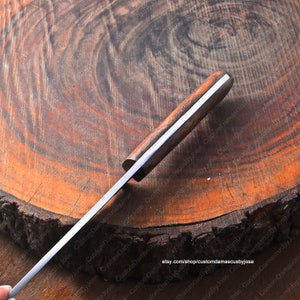 May include: A close-up of a long, thin, silver knife with a wooden handle. The knife is resting on a wooden surface with a circular grain pattern. The handle is made of dark wood with a light wood inlay. The knife is sharp and has a pointed tip.