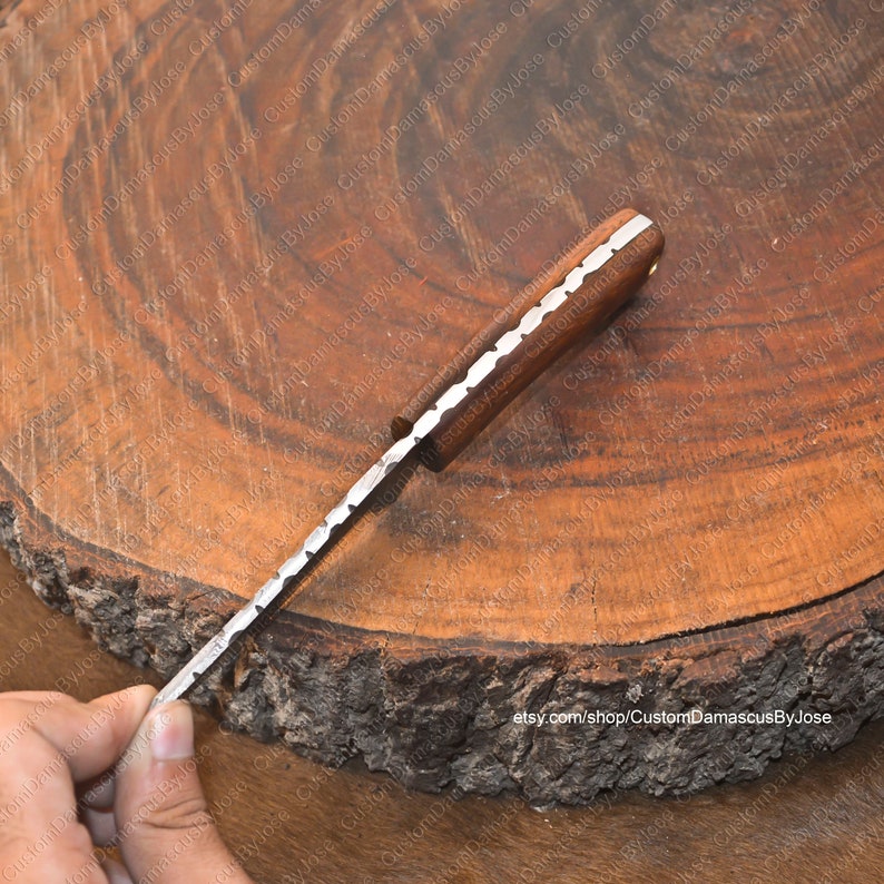 May include: A close-up of a handmade Damascus steel knife with a wooden handle. The blade has a wavy pattern and the handle is a dark brown color. The knife is resting on a wooden surface.