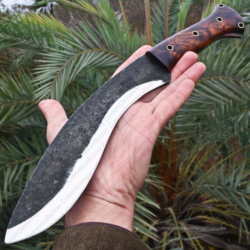 Puede incluir: Un cuchillo grande y curvo con una hoja oscura y texturizada y un mango de madera. La hoja tiene un borde blanco y un acabado oscuro y r&uacute;stico. El mango es de un marr&oacute;n rico con un veteado.