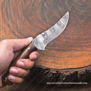May include: A close-up of a handmade Damascus steel knife with a wooden handle. The blade has a wavy pattern and is very sharp. The knife is resting on a wooden surface.