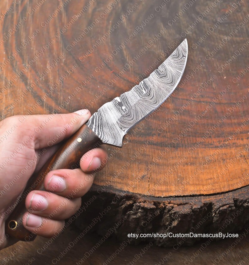 May include: A close-up of a handmade Damascus steel knife with a wooden handle. The blade has a wavy pattern and is very sharp. The knife is resting on a wooden surface.