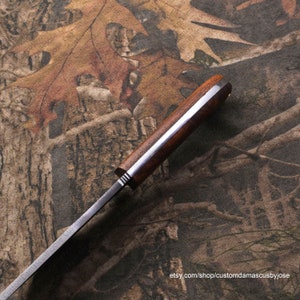 Puede incluir: Primer plano del mango de un cuchillo con mango de madera y hoja de acero. El mango es de color marr&oacute;n oscuro y tiene un acento de metal plateado. La hoja es de color plateado y tiene una ligera curva. El cuchillo est&aacute; apoyado sobre una superficie con estampado de camuflaje.