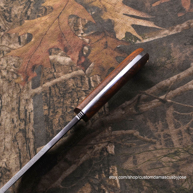 Puede incluir: Primer plano del mango de un cuchillo con mango de madera y hoja de acero. El mango es de color marr&oacute;n oscuro y tiene un acento de metal plateado. La hoja es de color plateado y tiene una ligera curva. El cuchillo est&aacute; apoyado sobre una superficie con estampado de camuflaje.