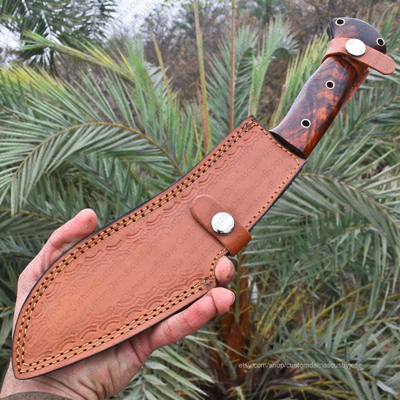 Puede incluir: Un cuchillo de caza hecho a medida con un mango de madera marr&oacute;n y una funda de cuero. El cuchillo tiene una hoja oscura y est&aacute; asegurado en una funda de cuero marr&oacute;n con un cierre de bot&oacute;n a presi&oacute;n. La funda tiene un patr&oacute;n en relieve.