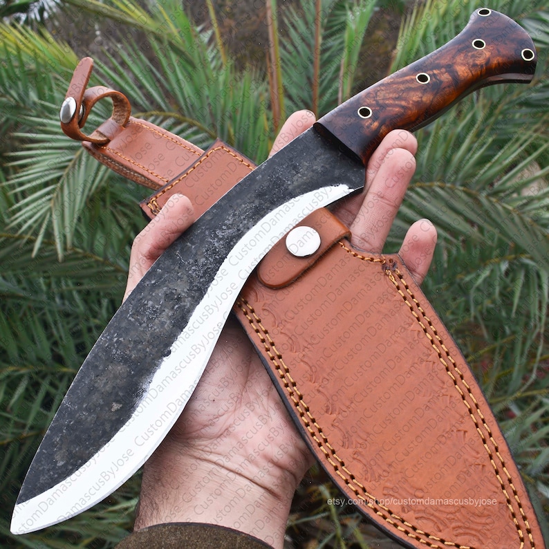 Puede incluir: Un cuchillo grande, artesanal, con una hoja oscura y texturizada y un mango de madera. El mango presenta una rica veta de madera marr&oacute;n con detalles en lat&oacute;n. Se combina con una funda de cuero marr&oacute;n con costuras decorativas.