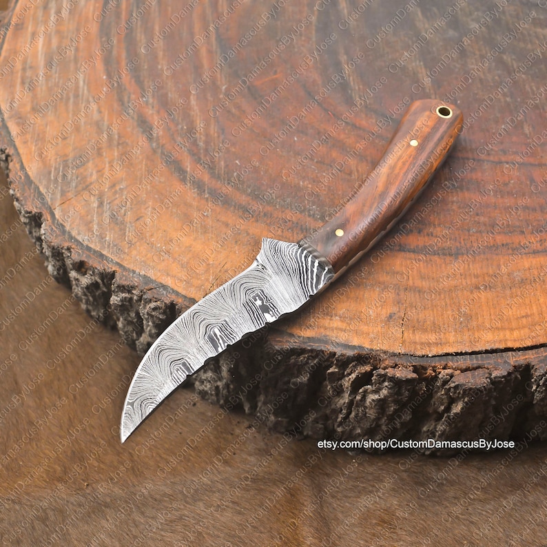 May include: A curved, Damascus steel knife with a wooden handle. The blade has a wavy pattern and is very sharp. The handle is made of dark wood with a light brown accent. The knife is resting on a wooden surface.