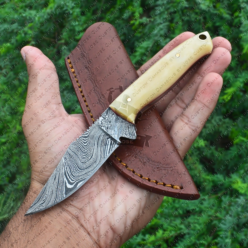 May include: A small, handcrafted knife with a damascus steel blade and a bone handle. The knife is in a brown leather sheath.