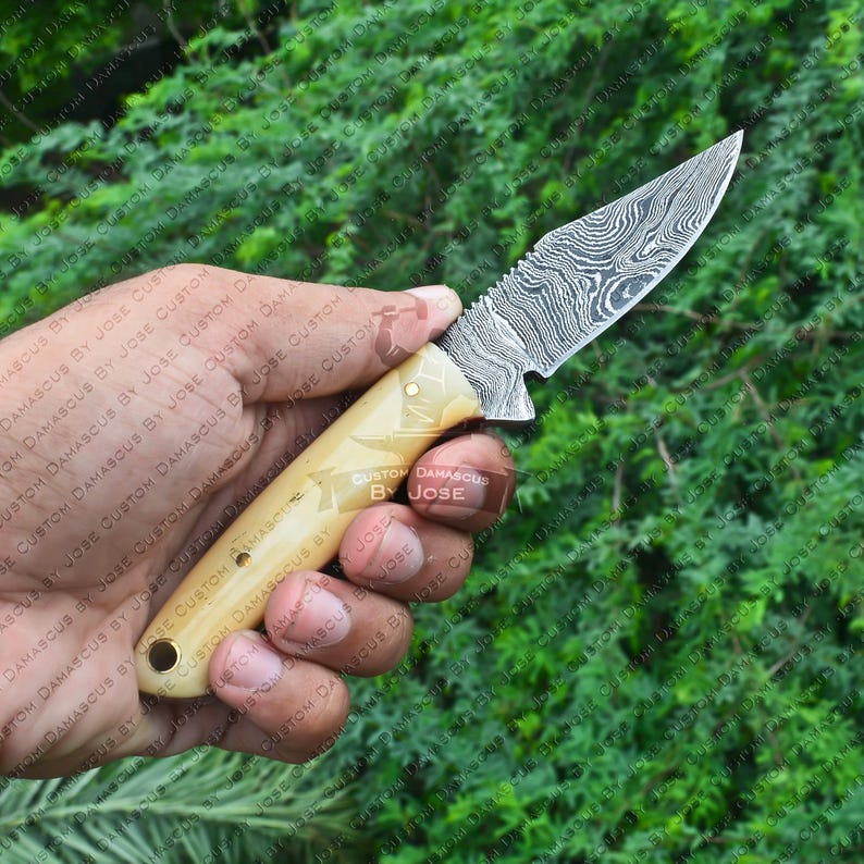 May include: A small, handcrafted knife with a patterned, stainless steel blade and a bone handle. The handle is decorated with the words "Custom Damascus by Jose".
