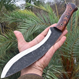 Puede incluir: Un cuchillo artesanal con una hoja oscura y curvada y un mango de madera. El mango presenta una rica veta de madera marr&oacute;n con detalles met&aacute;licos decorativos. La hoja tiene un borde blanco. El cuchillo se sostiene en una mano.
