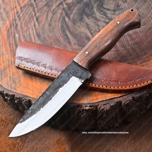 May include: A handmade knife with a dark wood handle and a leather sheath. The blade is made of Damascus steel and has a slight curve. The knife is resting on a wooden surface.
