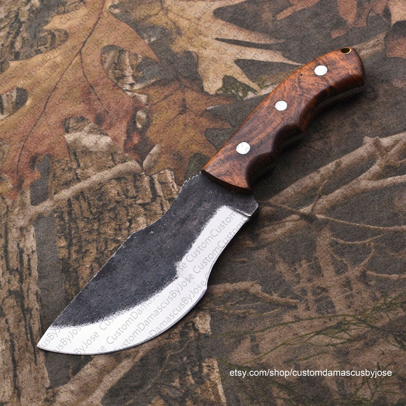 Puede incluir: Un cuchillo hecho a mano con una hoja curva y un mango de madera marr&oacute;n. La hoja est&aacute; hecha de acero Damasco y tiene un acabado pulido. El mango tiene una superficie texturizada y est&aacute; asegurado a la hoja con tres remaches de lat&oacute;n. El cuchillo est&aacute; apoyado sobre una tela con estampado de camuflaje.