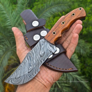 May include: A handmade Damascus steel knife with a wood handle and a leather sheath. The knife has a wavy pattern and a curved blade.