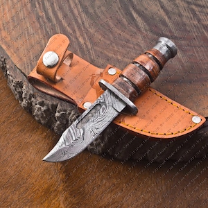 May include: A Damascus steel hunting knife with a wooden handle and a leather sheath. The blade has a unique pattern, and the handle is made of layered wood. The sheath is brown with a snap closure.