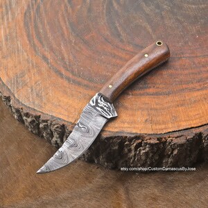 May include: A small, hand-crafted knife with a Damascus steel blade and a wooden handle. The blade has a wavy pattern and the handle has a brass pin. The knife is resting on a wooden surface.