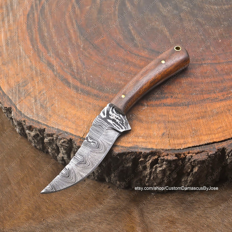 May include: A small, hand-crafted knife with a Damascus steel blade and a wooden handle. The blade has a wavy pattern and the handle has a brass pin. The knife is resting on a wooden surface.