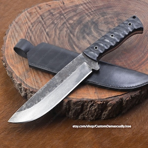 May include: A black-handled knife with a long, curved blade and a leather sheath. The knife is made of Damascus steel and has a textured handle. The knife is resting on a wooden surface.