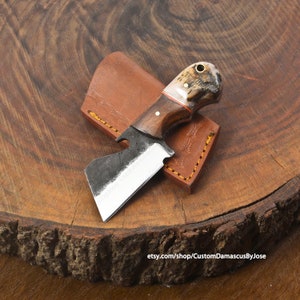 Pode incluir: Uma pequena faca feita à mão com um cabo de madeira castanho e uma virola de chifre de veado. A lâmina é de aço e tem uma ligeira curvatura. A faca está sobre uma superfície de madeira com uma bainha de couro.