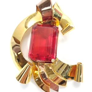 May include: Vintage gold-tone brooch with a large rectangular red gemstone center. The brooch features a ribbon-like design with curved and folded metal elements. A classic piece of jewelry.