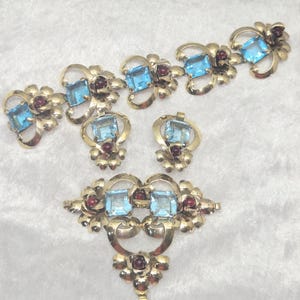 May include: A gold-toned jewelry set consisting of a link bracelet, a pair of earrings, and a large brooch. Each piece features square-cut light blue stones and small round dark red stones set within ornate, swirling gold-colored metal floral motifs.
