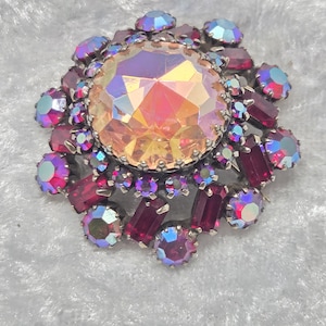 May include: A circular brooch featuring a large, central, faceted peach-colored stone with an iridescent finish. The center stone is surrounded by a cluster of smaller, deep red rectangular and round iridescent stones set in a silver-toned metal frame.