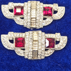 May include: Two Art Deco style brooches. Each brooch features a central rectangular section with clear rhinestones and a square, ruby-red stone on each end. The brooches are set on a silver-toned metal base and are displayed on a blue background.
