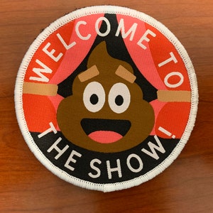 May include: A round, white-edged patch with a black background. The patch features a brown poop emoji with a smiling face and the text "Welcome to the Show!" in white.