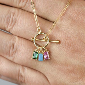 May include: Gold-coloured necklace with a toggle clasp and a chain link design. The necklace features three small, rectangular charms in green, blue, and pink. The necklace is displayed against a neutral background.