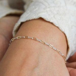 May include: A delicate bracelet featuring a repeating pattern of alternating silver and gold-toned rectangular chain links. The thin, minimalist chain is designed with a polished finish, creating a subtle contrast between the two metallic colors.